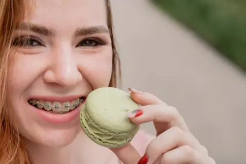 Uśmiechnięta dziewczyna z aparatem ortodontycznym sięga po zielonego makaronika. Kiedy można normalnie jeść po założeniu aparatu? Delikatne przysmaki są bezpieczne!