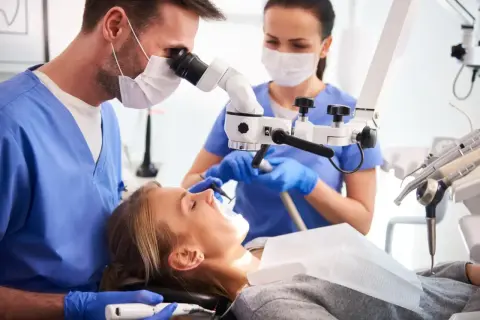 Dentysta pod mikroskopem bada reendo zęba pacjentki. Asystentka przygotowuje narzędzia.