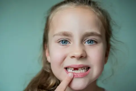 Uśmiechnięta dziewczynka z niebieskimi oczami pokazuje lukę po wypadniętym mleczaku. To znak, że czas na nowe zęby!