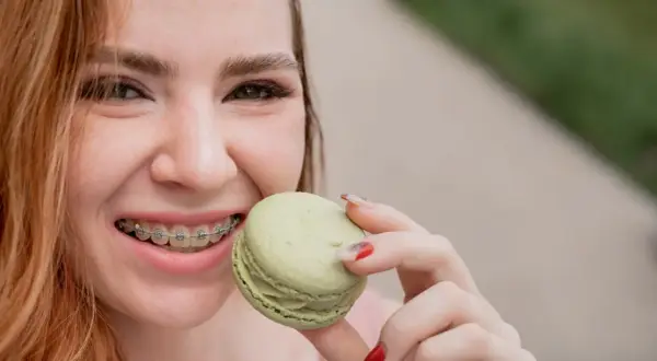 Uśmiechnięta dziewczyna z aparatem ortodontycznym sięga po zielonego makaronika. Kiedy można normalnie jeść po założeniu aparatu? Delikatne przysmaki są bezpieczne!