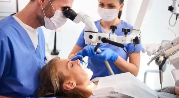 Dentysta pod mikroskopem bada reendo zęba pacjentki. Asystentka przygotowuje narzędzia.